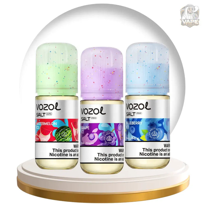 Three Vozol Salt e-liquid bottles in Watermelon, Grape, and Blueberry flavors, displayed side-by-side on a gold-rimmed white pedestal inside a clear glass dome. The bottles feature colorful speckled caps and vibrant fruit-themed labels.