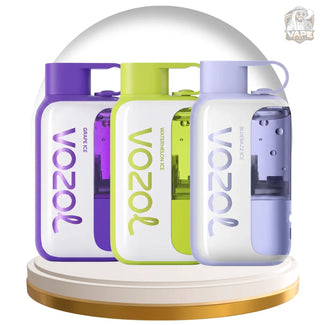 Three Vozol disposable vape devices in Grape Ice, Watermelon Ice, and Blue Ice flavors, displayed side-by-side on a gold and white circular pedestal. The devices feature white bodies with purple, green, and light blue accents, and are topped with a clear dome under a white studio background.