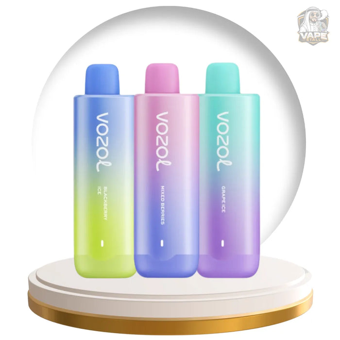 Three Vozol disposable vape devices in a row, each with a different pastel gradient color: blue to green, pink to purple, and teal to purple. They are displayed on a round white and gold pedestal with a white background and a Vozol logo in the top right corner.